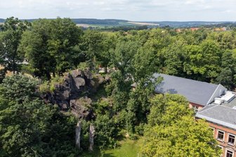 Gefahr in Falkenstein: Felsen droht Richtung Schule zu stürzen - Teile des Schulfelsens (links) könnten nach Ansicht von Experten in Richtung Falkensteiner Grundschule (rechts) stürzen. Ein Büro wurde jetzt mit der Planung von Sicherungsarbeiten beauftragt. Gesteinsbrocken könnten auf dem Grünstreifen (Mitte unten) mit einem Steinfallnetz gestoppt werden.