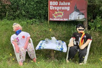Hier im Erzgebirge sind die Puppen los: So bereitet sich ein Dorf auf seine 800-Jahr-Feier vor - Am Ortseingang von Ursprung verdeutlicht das Plakat hinter den zwei Festpuppen, warum diese dort sitzen.