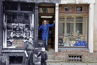 Hohenstein-Ernstthal: Wie lange gibt es den Tante-Emma-Laden von Ulrich Bohne noch? - Anna Bohne, die Frau des Gründers Alfred, mit einem ihrer Söhne vor dem Schokoladengeschäft (schwarz-weiß). Der heutige Inhaber Ulrich Bohne im historischen Eingang. Das Geschäft wurde 1930 um die Fläche hinter dem farbigen Schaufenster erweitert.