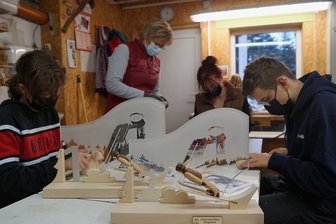 Klingenthaler Firma fertigt die Siegerpokale für den bevorstehenden Wintersport-Weltcup - Handgeschnitzte Skispringer vor der Silhouette der Vogtland-Arena: Die Pokale der Firma Holzkunst Müller aus Klingenthal für das Weltcup-Skispringen stehen bereit. Im Hintergrund Susanne Müller (stehend) mit Mia (links), Johannes und Kim von einer ihrer Schnitzkursgruppen. 