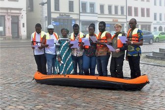 Kunstaktion: "Ode an die Freude" erklingt im Schlauchboot auf dem Freiberger Obermarkt - Sechs Studenten aus Ghana und Nigeria, verstärkt von Dozentin Ramatu Leeder-Kamanda (3. v.l.) und Stadträtin Jana Pinka (Die Linke, 7. v. l.), sangen auf dem Obermarkt in Freiberg die Ode an die Freude. Die Aktion des Konzeptkünstlers Shannon Alexander Murphy soll für Hoffnung und Verständigung stehen.