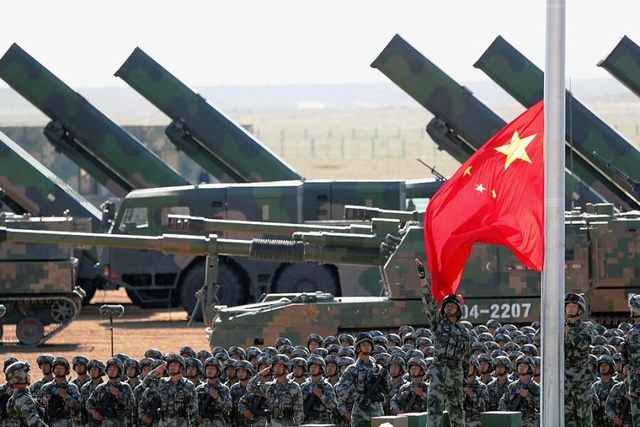 Militärexperte zu Chinas Aufrüstung: "Entwicklungen in China sind besorgniserrgend für die USA" - Zur Militärparade zum 90. Geburtstag der Volksbefreiungsarmee auf der Zhurihe Trainings-Basis, 400 Kilometer nordwestlich von Peking, im Jahr 2017 wurde eine Fahne gehisst. Im Hintergrund sind Haubitzen und Raketenwerfer zu sehen. 
