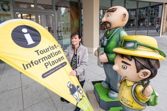 Modernisierung der Plauener Tourist-Information beginnt: Was Einwohner und Besucher der Stadt jetzt wissen müssen - Kathleen Dentler von der Tourist-Information zeigt, wo es langgeht: Ein Aufsteller sowie die Figuren von Vater und Sohn weisen den Weg zum Ausweichquartier im Rathausfoyer.