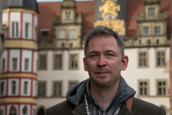 Name, Arbeit, Wohnen – was macht den typischen Zwickauer aus? - Das ist Thomas (48) aus Zwickau – zumindest nach den Vorstellungen eines Bildprogramms, das Künstliche Intelligenz (KI) einsetzt. Im Hintergrund sieht man das von der KI erschaffene Zwickauer Rathaus. Es ähnelt dem echten nur wenig.