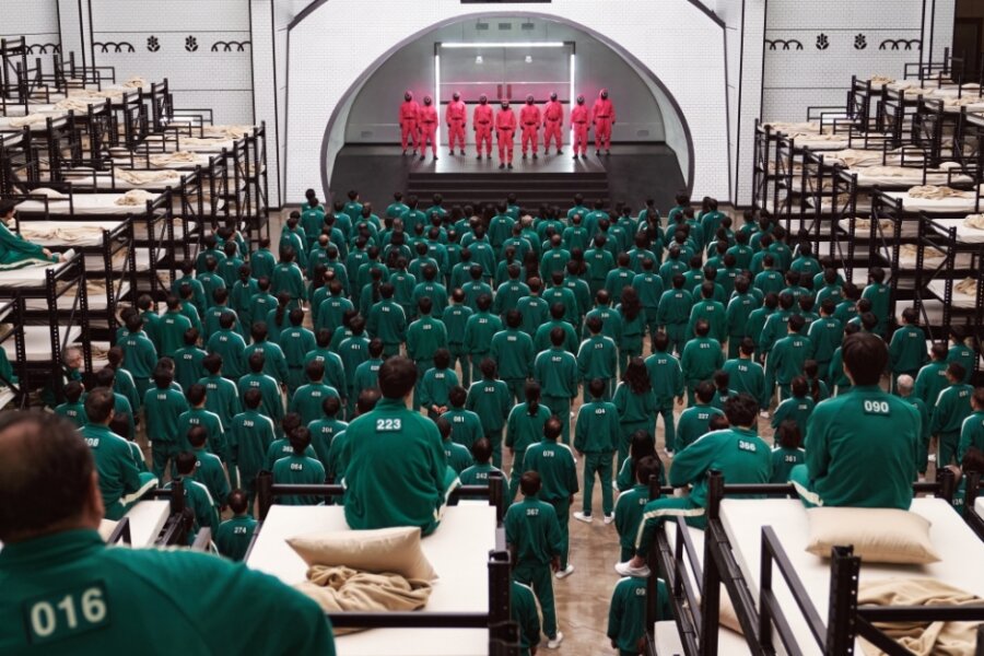 Netflix-Serie "Squid Game" weckt in Zwickau Interesse an Korea - Die Antagonisten in "Squid Game": Die Teilnehmer des Spiels in dunkelgrünen nummerierten Trainingsanzügen, die Wächter in rote Kapuzen-Overalls und schwarzen Fechtmasken.
