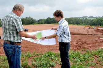 Oelsnitz/Gewerbegebiet Hoffeld: Zentraler Fußballplatz nimmt Gestalt an - Am Gewerbegebiet Hoffeld entstehen auf einem rund 24.000 Quadratmeter großen städtischen Grundstück ein Kunstrasenplatz mit 90 mal 60 Meter großem Spielfeld (links im Bild) und ein 105 mal 68 Meter großer Rasenplatz (rechts) sowie ein Funktionsgebäude. Links sind auch die Betonelemente für die Terrasse zu erkennen. Holger Klier (links) und Thomas Illmann stehen auf der Halde aus Mutterboden.