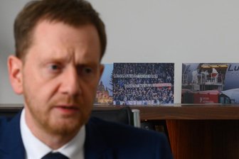 Offener Brief an sächsischen Ministerpräsidenten: 230 Unterschriften gegen rechts - Ministerpräsident von Sachsen sitzt bei einem Interview mit der Deutschen Presse-Agentur in seinem Büro in der Sächsischen Staatskanzlei vor aufgestellten Fotos mit Szenen der Corona-Pandemie. Unter anderen ein Foto des Banners "Kretschmer, du willst Sachse sein? Verhältst dich wie ein Wessischwein!", das beim Zweitligaspiel am 7. November 2021 in Aue von Fans gezeigt wurde.