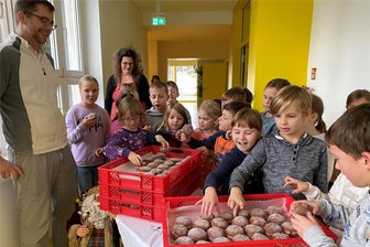Pfannkuchen für alle: Eine Schülerin der Grundschule Hauptmannsgrün teilt ihr Tombola-Glück mit Schülern und Lehrern - Nicole Spiller und Robert Hartmann von der Bäckerei Zeidler lösten mit ihrem Besuch in der Grundschule Hauptmannsgrün große Freude aus - zwischen den beiden lässt sich‘s Meggie Mollenhauer schmecken, die Gewinnerin der Pfannkuchen.