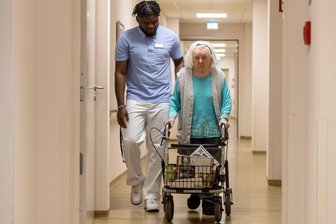 Pflegenotstand: Hartmannsdorf setzt auf internationale Talente - Bertister Dzuffo Meli begleitet Magdalena Birkholz durch das Azurit-Seniorenzentrum in Hartmannsdorf. Die 93-Jährige ist auf Hilfe beim Gehen angewiesen.