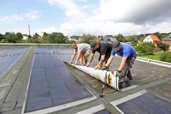 Sanierer steigen Schwimmern im Hot-Badeland aufs Dach - Mit purer Muskelkraft, aber vorsichtig lösen die Dachdecker die aufgeklebten Fotovoltaiksegmente von der Oberfläche des HOT-Badeland-Daches. Das hat Risse bekommen, durch die Wasser in großen Mengen eindringen konnte.
