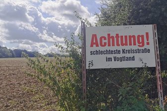 "Schlechteste Kreisstraße im Vogtland": Kein Baustart in Sicht - An der K 7884, etwas zurückgesetzt, steht diese Tafel als Mahnung an Autofahrer, die sich auf die desolate Kreisstraße trauen. Die geflickte, bucklige Fahrbahn bröselt, Bankette sind teils nicht mehr vorhanden.