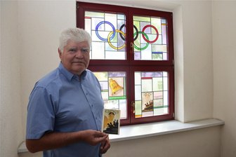 Sensationelle Entdeckung: Die seit 90 Jahren unbemerkten Olympia-Fenster von Zwickau - Sporthistoriker Werner Beuschel mit dem Olympia-Plakat von Berlin 1936 vor dem Bleiglasfenster, in dem sich das gleiche Motiv befindet.