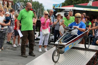 Syrauer Dorfstraße wird zur Rennpiste: Ex-Bürgermeister steigt auf Rasentraktor - Frank Wunderlich vom Männergesangsverein Syrau wurde Zweiter. Pfarrer Michael Kreßler (links, mit Mikrofon) verstand es, mit viel Witz und Schlagfertigkeit die einzelnen Starter anzukündigen. 