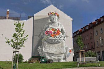 Tassos Marktfrau in Meerane: Warum Stadttechnik-Mitarbeiter mit einer Hebebühne am Wandbild anrücken - Das Motiv der Marktfrau ziert eine Fassade in der Innenstadt von Meerane. Am Wandbild ist ein Einsatz der Stadttechnik geplant.