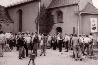 Themen ändern sich - Idee bleibt - Teilnehmer des Friedensseminars in den 80er-Jahren vor der Kirche Königswalde, rechts an der Fassade das Symbol des Treffens, der umgedrehte DDR-Stahlhelm aus dem eine Sonnenblume wächst.