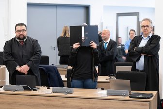 Urteil gegen Lina E. gefallen: Mehrjährige Haftstrafe - Die Angeklagte (M) steht bei der Fortsetzung des Prozesses im Oberlandesgericht (OLG) Dresden im Verhandlungssaal und hält einen Aktenordner vor ihr Gesicht. Die Gruppe bestehend aus Lina E. und drei Männern aus der linksextremistischen Leipziger Szene ist wegen Mitgliedschaft in einer kriminellen Vereinigung angeklagt. Foto: Sebastian Kahnert/dpa