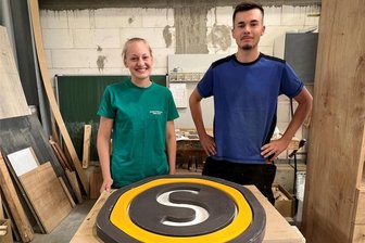 Wo in Frankenberg die Tischler von morgen lernen - Die Tischlerlehrlinge Helen Haase und Jonas Behm mit dem neuen Logo des Unternehmens Sachsenholz.